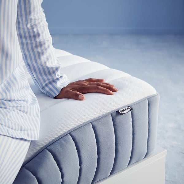 Person sitting on white MALM bed mattress, light blue ikea tag visible, against blue wall.
