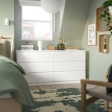 MALM chest against green wall with books, lamp, vase. Tall, matches nearby chairs. Park print right, window above.