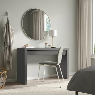 Modern grey MALM dressing table with mirror. Sleek glass top, single drawer, light-colored chair. Placed near bed and window, with coat hooks nearby.