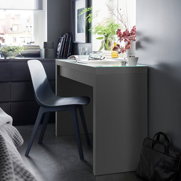 Modern grey MALM desk with glass top, beside a window, chair tucked. Potted plant, books, lamp. Minimalist design, functional storage.