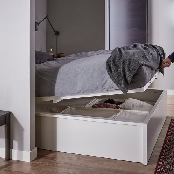 A white MALM storage bed with grey bedding. A compartment opens under the bed revealing folded clothes.