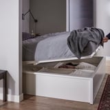 A white MALM storage bed with grey bedding. A compartment opens under the bed revealing folded clothes.