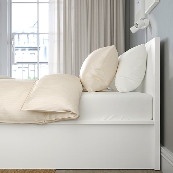 Minimalist bed with lift-up base for under-bed storage; large pillows, curtains, and window view.