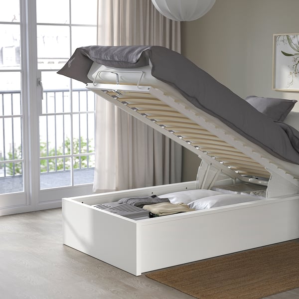 Bed with lifted storage base, showing inside space. Grey pillows and quilt on a light wooden floor, by a balcony window.