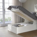 Bed with lifted storage base, showing inside space. Grey pillows and quilt on a light wooden floor, by a balcony window.