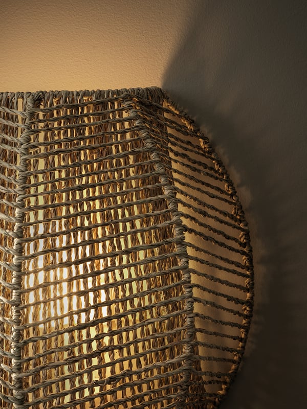 Hand-braided wall lamp, MÅNALG, emits warm light. Sedge material, brown-grey, hexagon shape, casts intricate shadows.