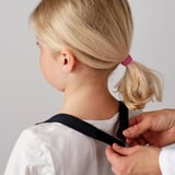Child wearing white apron with touch-and-close closure being fastened by adult. The apron protects clothes during kitchen activities.