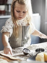 A young girl in a white apron with brown stripes is cooking, hands in flour. She wears a floral shirt. The apron protects her clothes while she bakes.