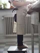 Child wearing MARIATHERES apron stands on stool, holding phone. Apron is beige, made of cotton and linen.
