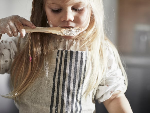 A young girl wears a white and gray striped kitchen apron, tasting batter with a wooden spoon.