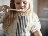 A young girl wears a white and gray striped kitchen apron, tasting batter with a wooden spoon.