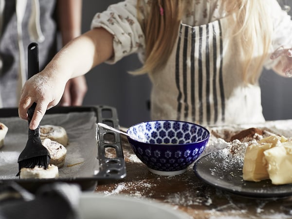 Person is baking, using a brush to apply liquid on pastries, with bowls and ingredients nearby. Apron worn, counter messy with flour.