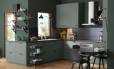 Modern green-grey kitchen with METOD cabinets, black fixtures, and black chairs. Shelves hold plants and cookware.