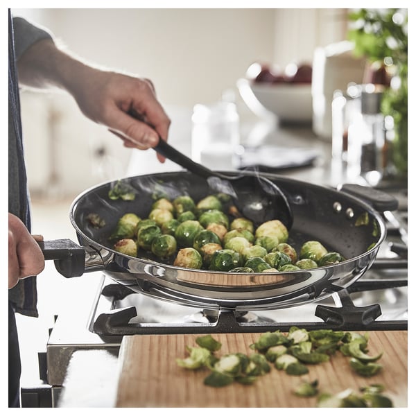 A person stirs brussels sprouts in a black MIDDAGSMAT frying pan on a gas stove. Wooden cutting board with sprouts nearby.