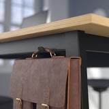 Brown leather bag hangs on black metal hook, linked to wooden shelf, fitting office decor.