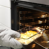 A hand holding a glass dish with baked food, removing it from an oven. The dish is oven-safe and can go directly to the table.