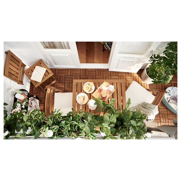 Cozy balcony set-up with NÄMMARÖ wood table and chairs, white cushions, green plants, and food on the table.