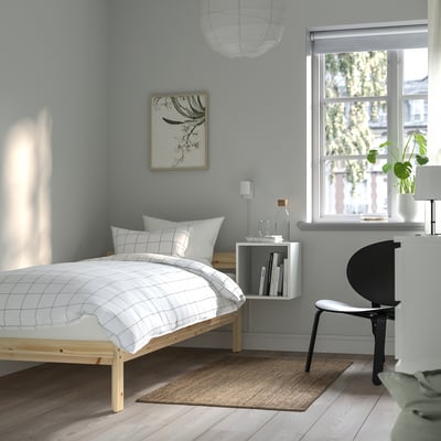 NEIDEN bed in natural wood, small size, low height, white duvet cover, minimalist design. Hanging artwork above. Chair and plant by window.