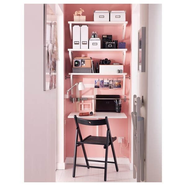 Compact home office setup with foldable black chair, pink walls, shelves with storage boxes, camera, laptop, and desk lamp.