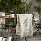 OLIVSPINNMAL throw blanket: white w/ orange, green patterns. Hangs on outdoor chair, larger than surrounding cups, between similar chairs & table.