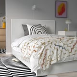 Small bed, white headboard, colorful blanket and rainbow pillow, smaller than nightstand, larger than slippers.