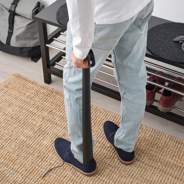 Person using long metal shoehorn while standing on woven mat.