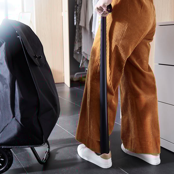 Person with brown striped pants and white sneakers holding a black shoehorn next to a black bag.