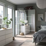 Modern bedroom with PAX wardrobe in white, mirror door, gray accents, wooden floor, plants, and minimalist decor.