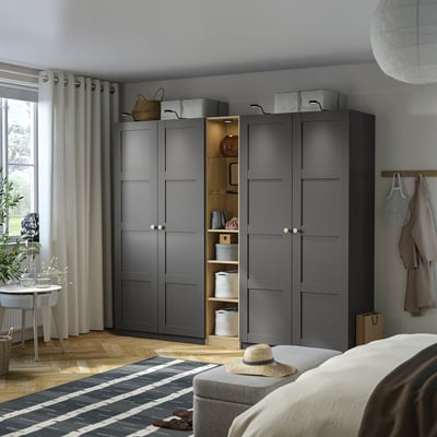 Black PAX wardrobe with shelves & drawers, beside curtain, table with plant, partially hiding couch with hat.