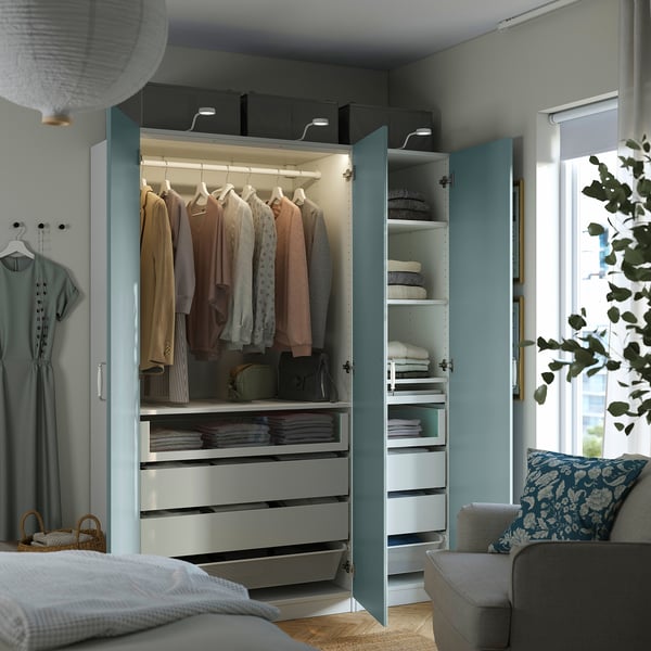 Modern bedroom with open wardrobe displaying folded clothes, drawers, and cushions, near a window with light blue decor.