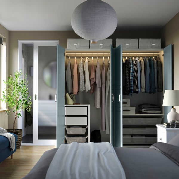 Bedroom with open wardrobe displaying clothes, drawers, shelves, and lighting. A lamp and plant are visible beside it.