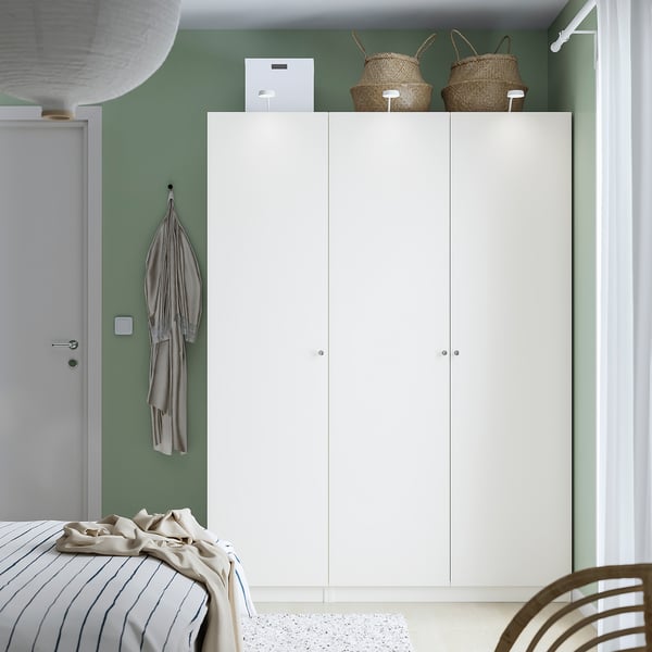 A modern bedroom with a large white PAX wardrobe and bed, decorated with neutral tones, green walls, and wicker baskets on top.