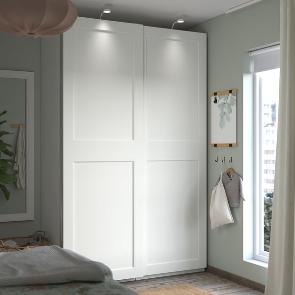 A white PAX wardrobe stands against a green wall. It has sliding doors with built-in lights, a hanging rail, and a mesh basket for storage.