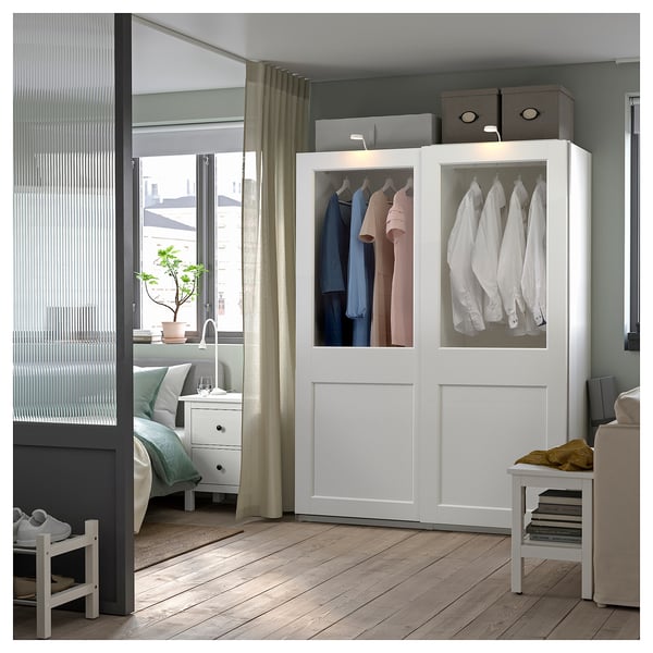 A modern white PAX wardrobe with glass top doors, displaying colorful clothes in a bedroom with wooden floors and natural light.