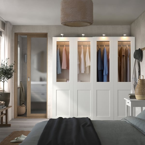 Modern bedroom with white PAX wardrobe, glass doors, colorful lamp, dark gray bed, wooden bench, and plant.