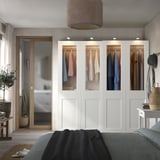 Modern bedroom with white PAX wardrobe, glass doors, colorful lamp, dark gray bed, wooden bench, and plant.