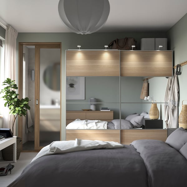 Sleek PAX wardrobe in light wood with mirrored doors, soft lighting, organised shelves, and a cosy bed with grey linens.