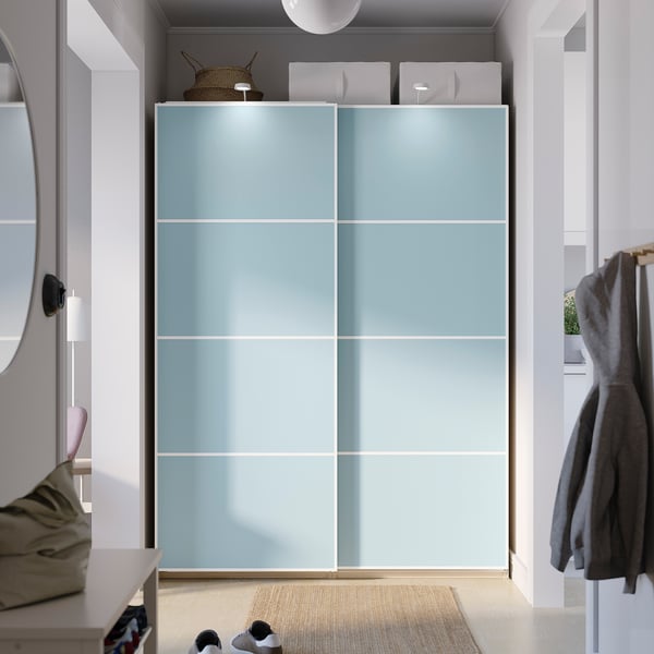 Modern light blue PAX wardrobe with sliding doors, mirror left, coat rack right. Dominates room with ample storage.