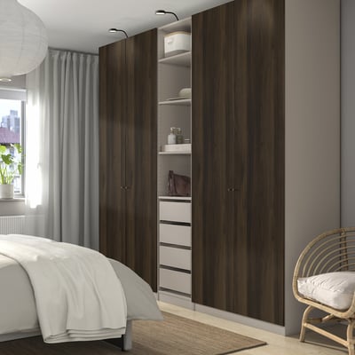 Modern bedroom with large dark wood PAX wardrobe, white bed, grey curtains, and wicker chair.