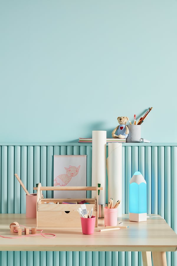 Kids desk with blue light, art supplies, teddy bear. Pink cups, wooden toys. Light blue walls, vertical lines.