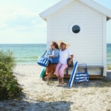 Two women sit by a beach hut, one holding a blue PILLERSTARR cooling bag larger than a handbag.