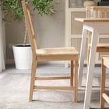 Light brown wooden chair with vertical slat back and simple design, placed near a white table with a plant in the background.