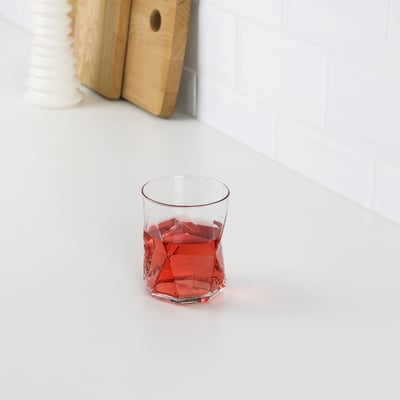 A red geometric glass cup on a white surface. The faceted design catches light, creating an elegant drink presentation.