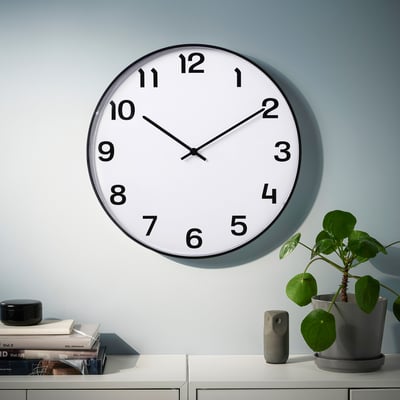 Wall clock, black PLUTTIS, quartz movement, silent, above shelf with plant & books.