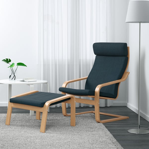 Dark blue POÄNG recliner with footrest, lamp, rug, side table, plant.