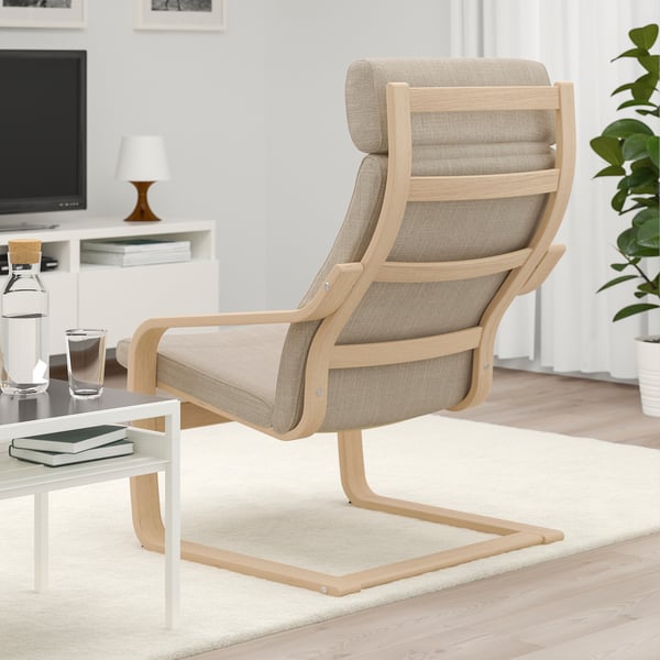 Modern living room with a beige POÄNG armchair, footstool, and coffee table. Books, water, and a tv in the background.