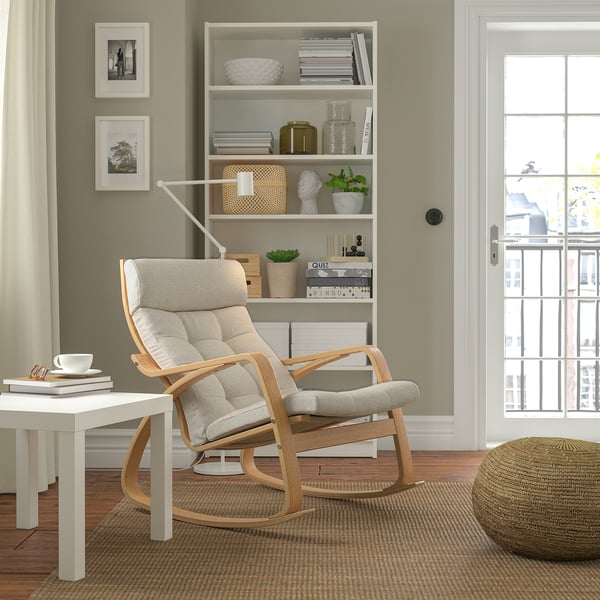 Rocking chair POÄNG with cushions on rug, next to table and book shelf.