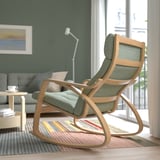 A modern rocking chair with a green cushion in a living room by a window. Its part of the POÄNG series, designed for comfort and durability.