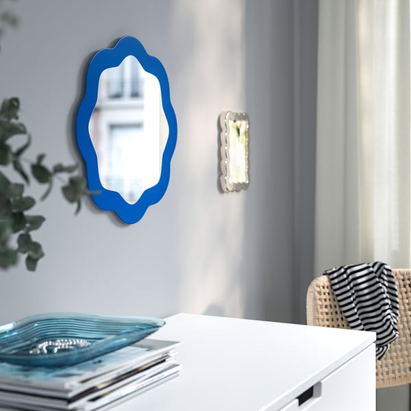 Blue-framed, wavy mirror on wall, reflecting interior. Beside white drawer with books and decorative tray.