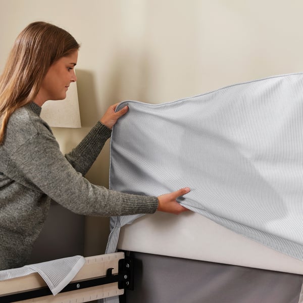 A woman is making up a bed with a striped RAMNEFJÄLL bed frame. She is smoothing the removable cover over the padded headboard.